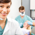 A portrait of a dental assistant smiling at the camera with the dentist working in the background.