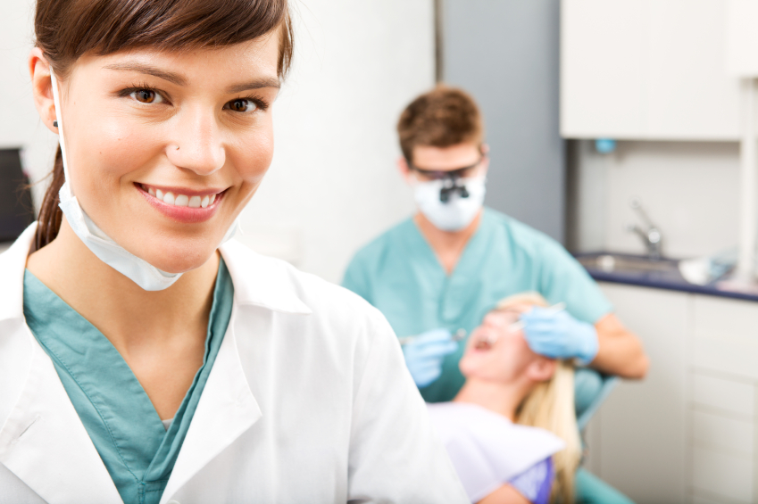 A portrait of a dental assistant smiling at the camera with the dentist working in the background.